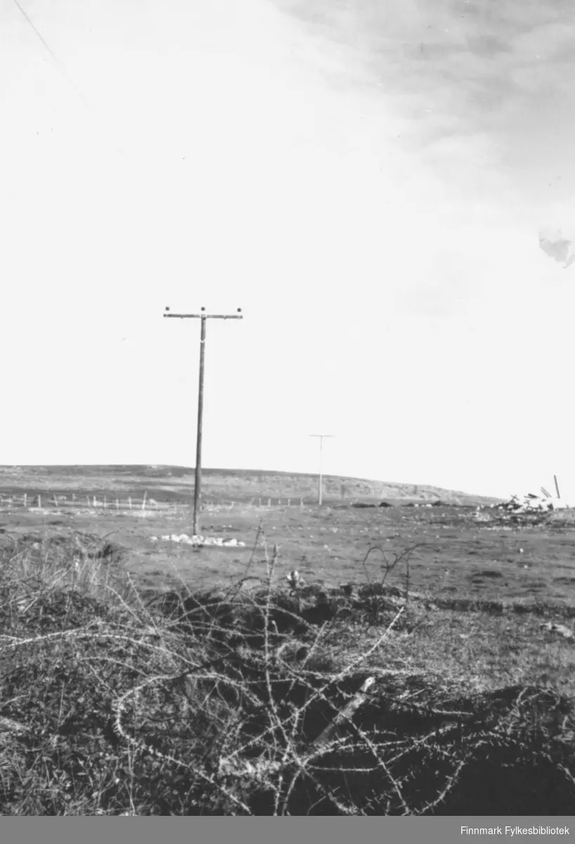 Landskapsmotiv fra Vadsø like etter andre verdenskrig. I forgrunnen ligger en haug med piggtråd. Fra venstre hjørne og på skrå innover i bildet ser man stolper og elektrisitetslinjer. I bakgrunnen er det fjell og åpent landskap. Bakerst i høyre hjørne ligger en ruinhaug
