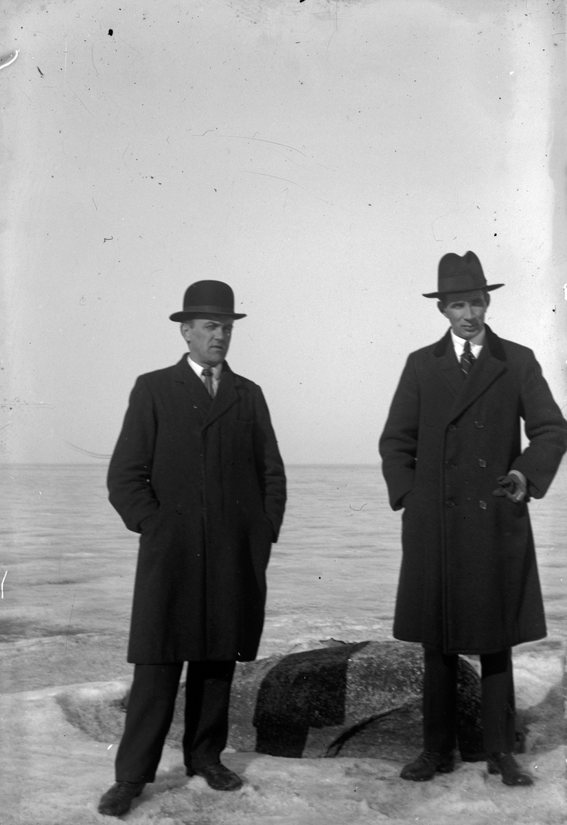 Två män i rock och hatt står vid en isbelagd sjö.