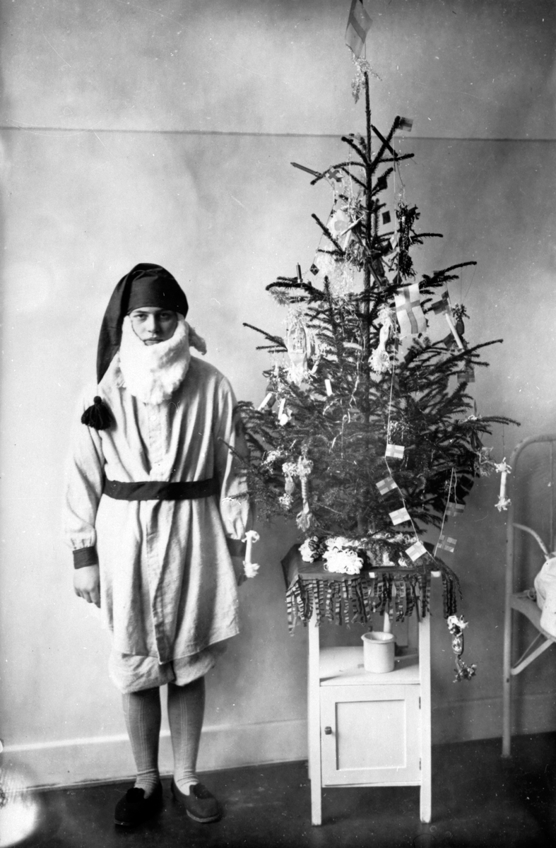 Julfirande vid Eksjö sanatorium år 1921. En pojke är utklädd till tomte. En liten gran, som är klädd med ljus och flaggor, är placerad på ett nattduksbord bredvid en sjuksäng i en sjuksal.