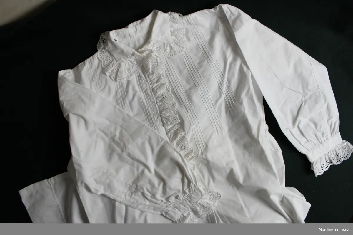 Hvit nattkjole for voksen. Knapper foran, nederste del hel. Krage med påsydd blondekant. Blondekant påsydd over knappedelen. Lange ermer med påsydd blondekant. Knapping. Bisesøm i forkant - pyntesøm. Hjemmelaget