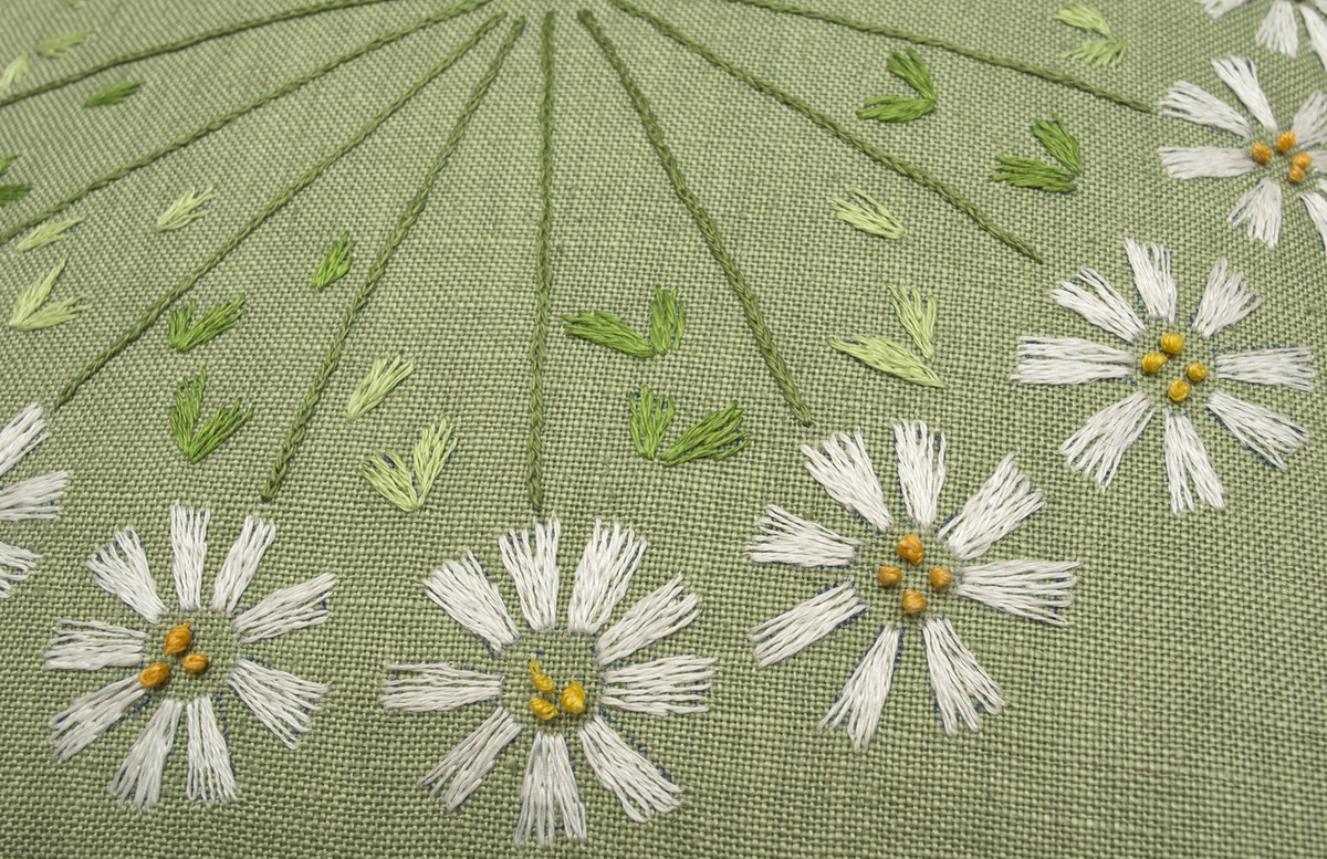 Stoppad kudde. Broderad på ljusgrönt linne (Ölandslinne färg 205) med klyvsöm, kedjesöm och knutsöm i vitt, gult och grönt lingarn. Baldersbrå-blommor på stjälk där blommorna bildar krans och stjälkarna strålar in mot mitten.