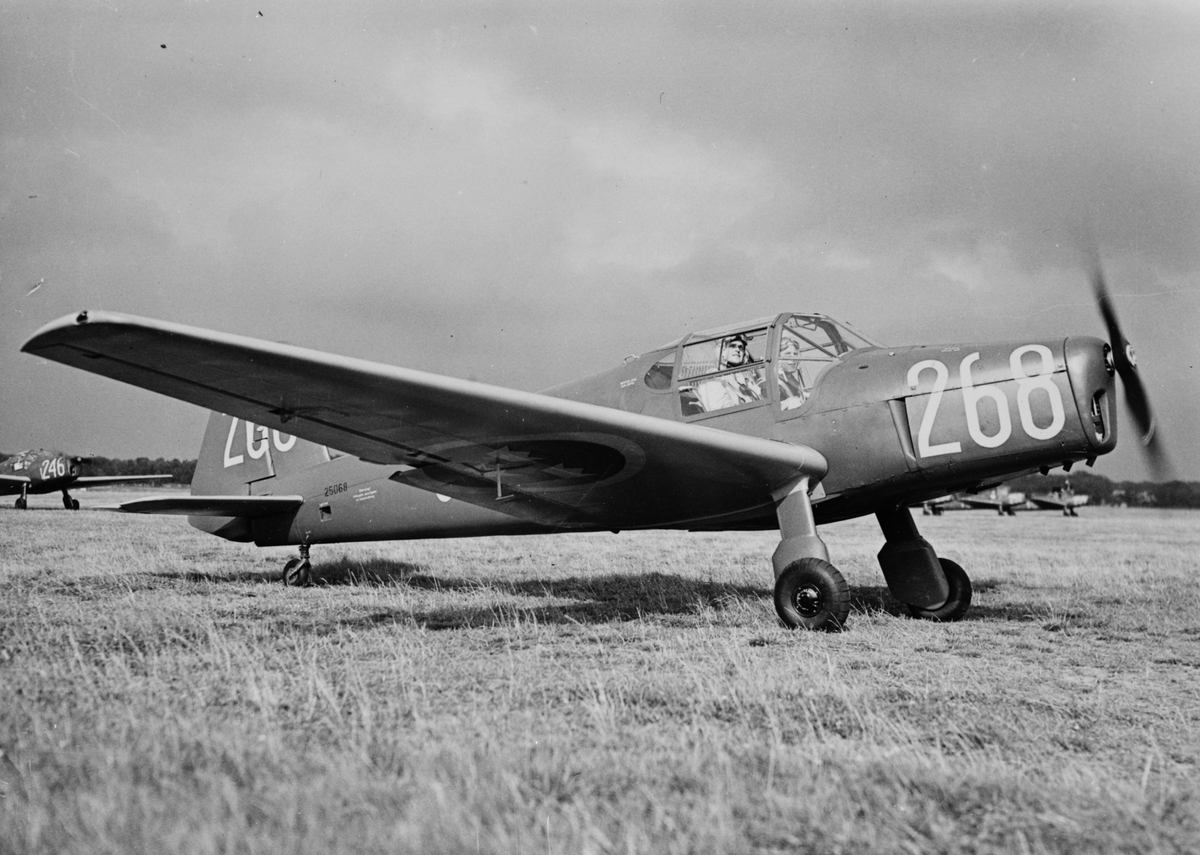Flygplan SK 25 märkt nummer 268 (25068) med flygförare och flygelev står på ett flygfält. I bakgrunden syns fler flygplan SK 25. Flygplan tillhörande Krigsflygskolan F 5.