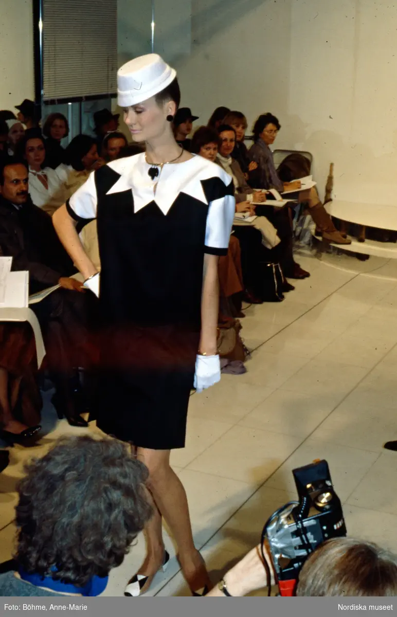 Modevisning. Modell i svart klänning med vit stjärnformad krage, vit hatt och smycken. Från Courrèges.