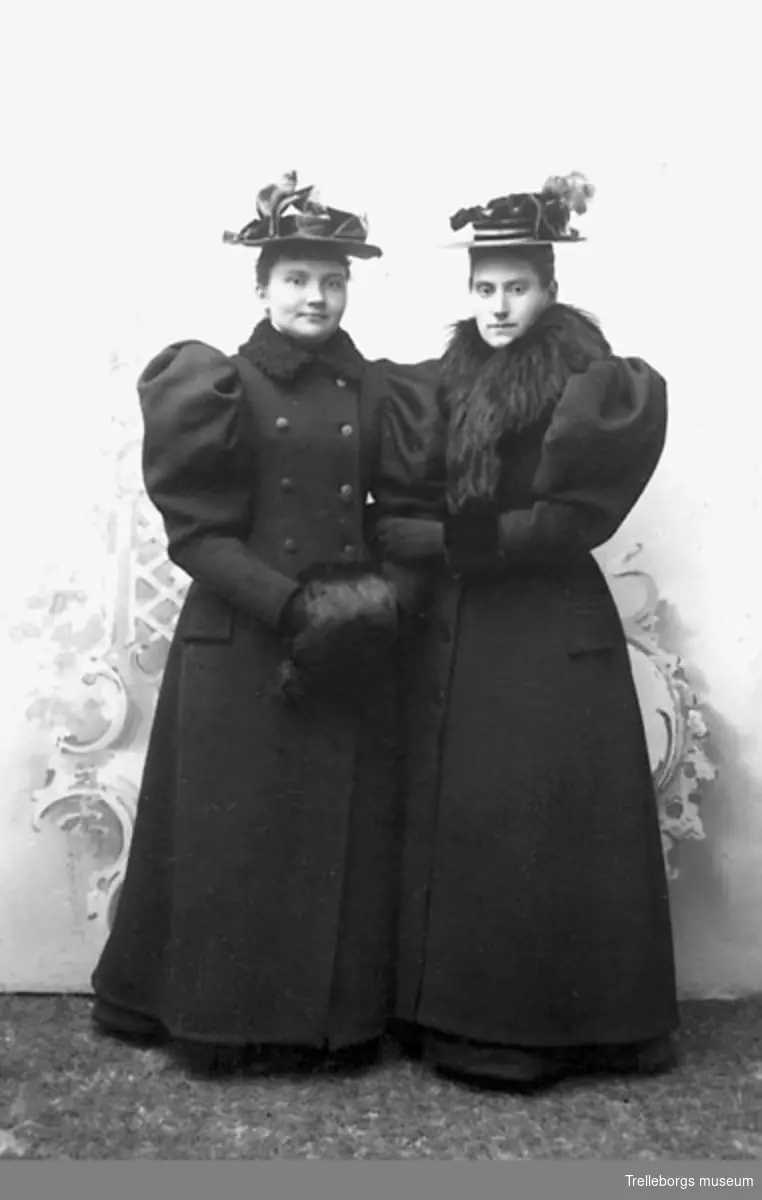 Två kvinnor i svart lång kappa med vida ärmar och pälskrage. En med pälsmuff. Båda i hatt.