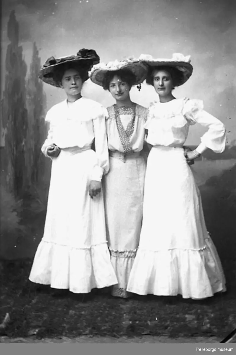 Tre kvinnor i sommarklänning med volang nertill och bredbrättad hatt.