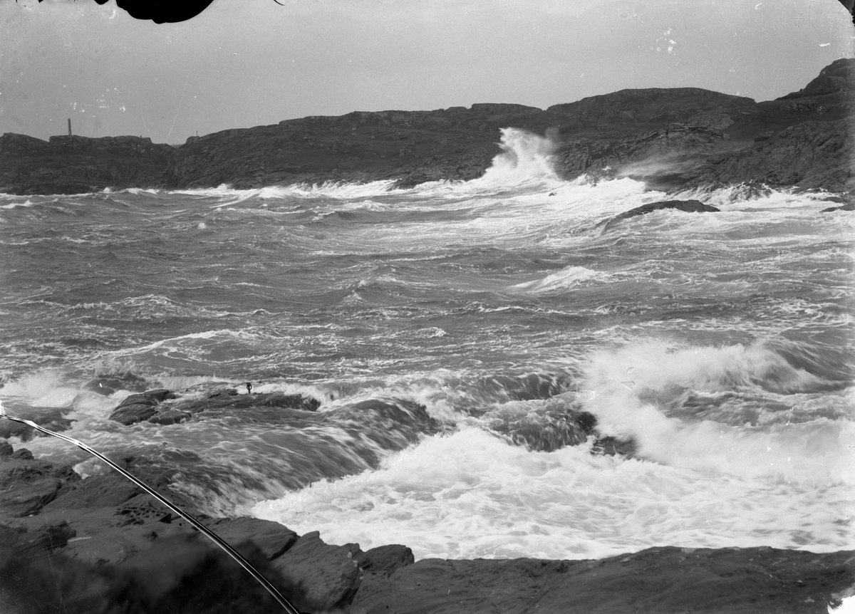 Sjø med bølger som slår inn mot land i Gardsvik (?). Fyrtårn til venstre (?) og steinkorset på Gard (?) (midt på bildet). Fjell i bakgrunnen.