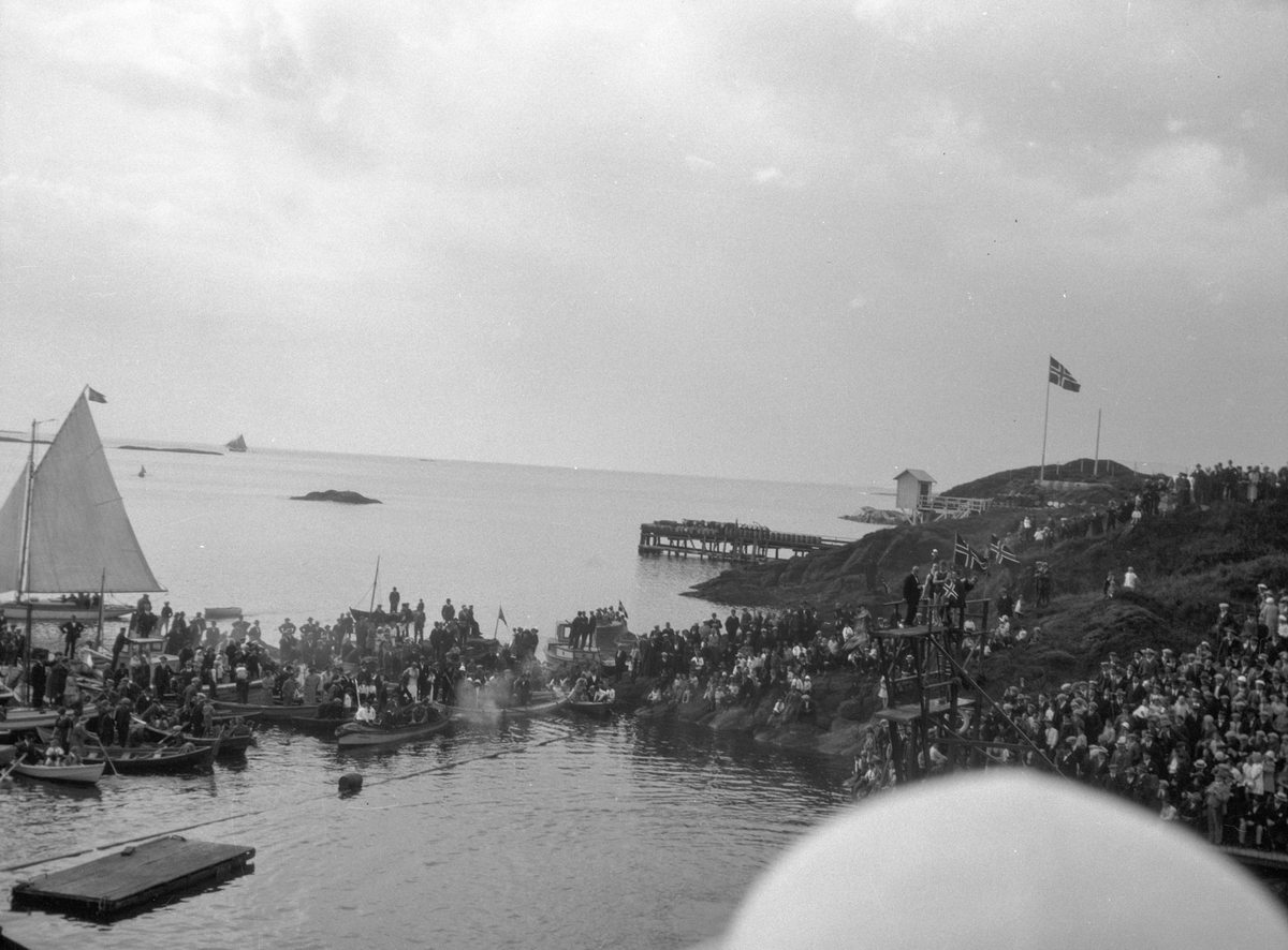 Sjøbadet i Asalviken.  Mange personer på land. Flere færinger og noen seilfartøyer på sjøen, med mange personer ombord. Brygge til høyre. Mindre brygge til venstre. En flaggstang og flere norsk flagg.