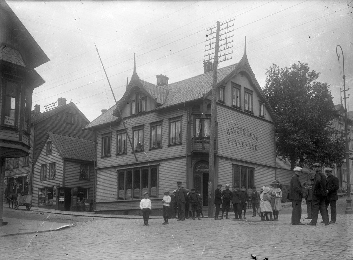 Haugesunds Sparebanks gamle bygning. Barn og voksne på gaten ved siden av. Trehus til venstre. Trær bak banken.