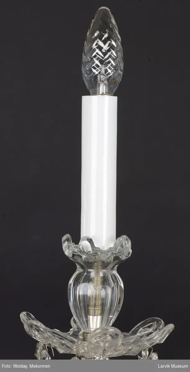 Balusterformet lysekrone.Slepen stamme med 6 svugne lyseholdere. Glassskål skjuler armfeste av stål, prismer i kjeder og enkle. Imitasjon av stearinlys i glass.
