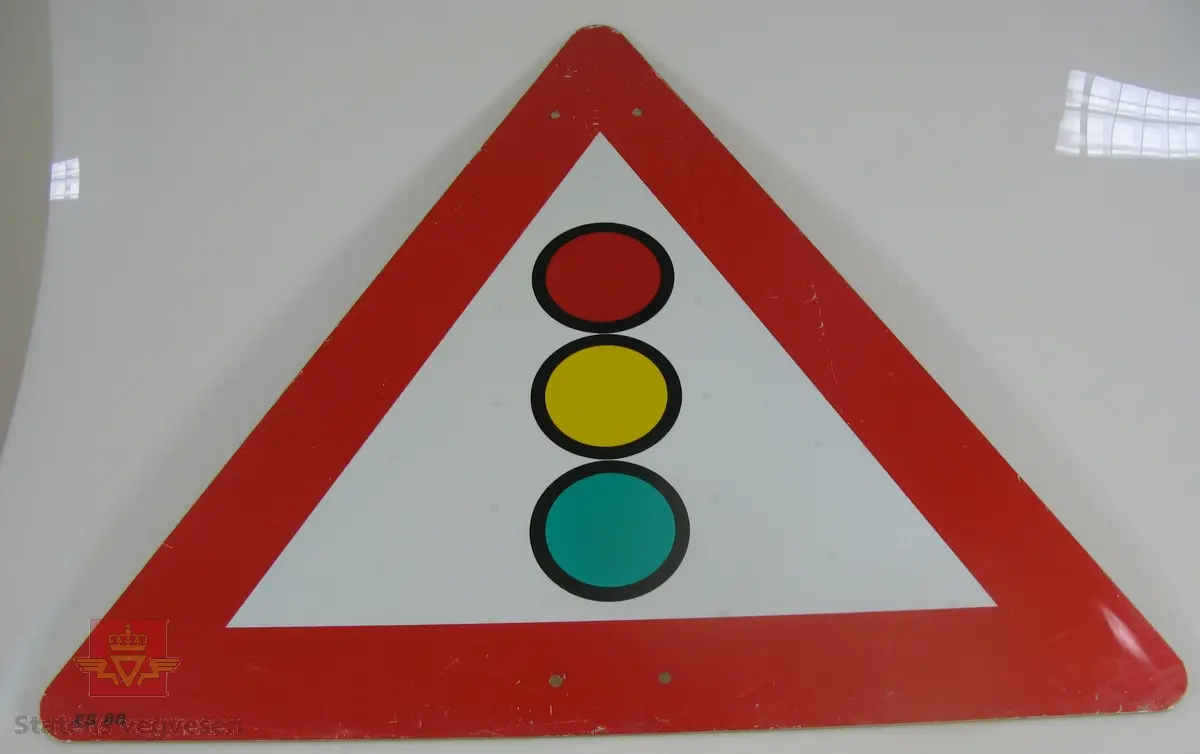 Trekantet skilt av tre mm aluminium, skiltet har avrundede hjørner. Rød bord og flerfarget symbol på hvit lysreflekterende bunn. Symbolet varsler om trafikklys. Grå bakside. Fire hull for fastskruing. Merking fra produsent.