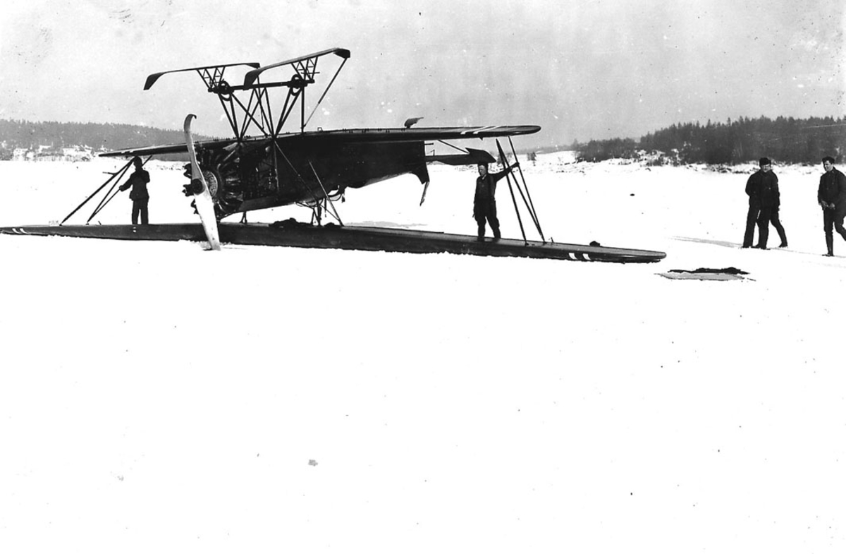 Åpen plass flere personer ved ett fly, Fokker CVE 311 med skiunderstell, snø på bakken. Flyet ligger på "rygg". Havarert 