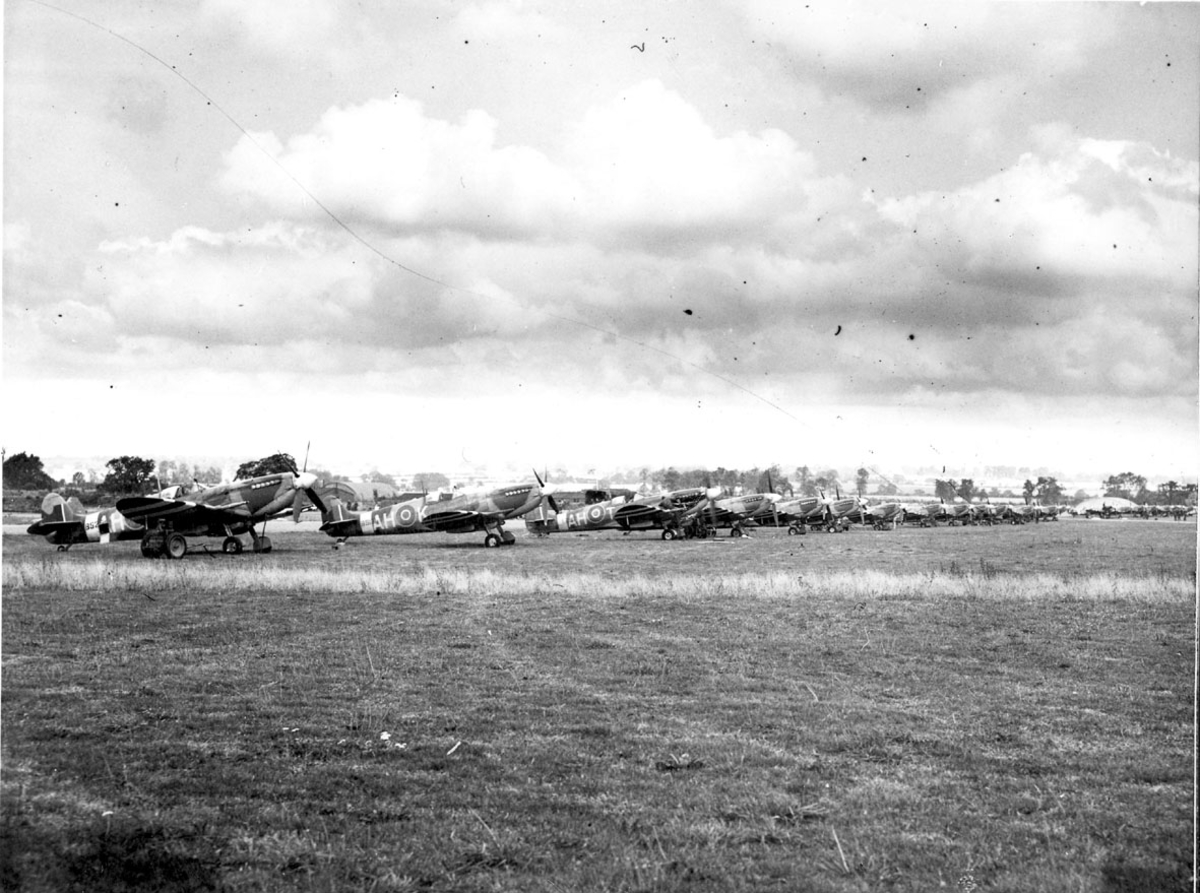 Åpen plass, ant. lufthavn, med mange fly oppstilt på rekke, Spitfire, skrått forfra.