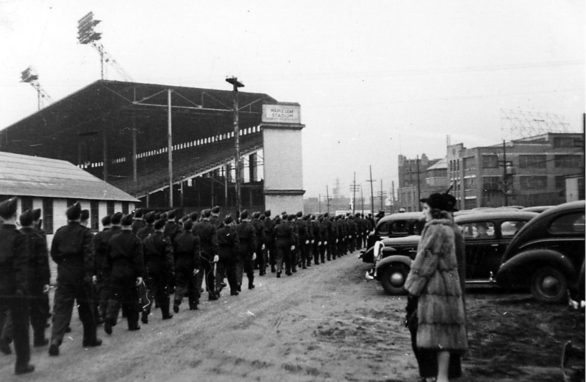 Gruppeportrett, lang rekke med soldater, militære, i militæruniform masjerer inn på et stadion. Noen biler og to kvinner t.h.