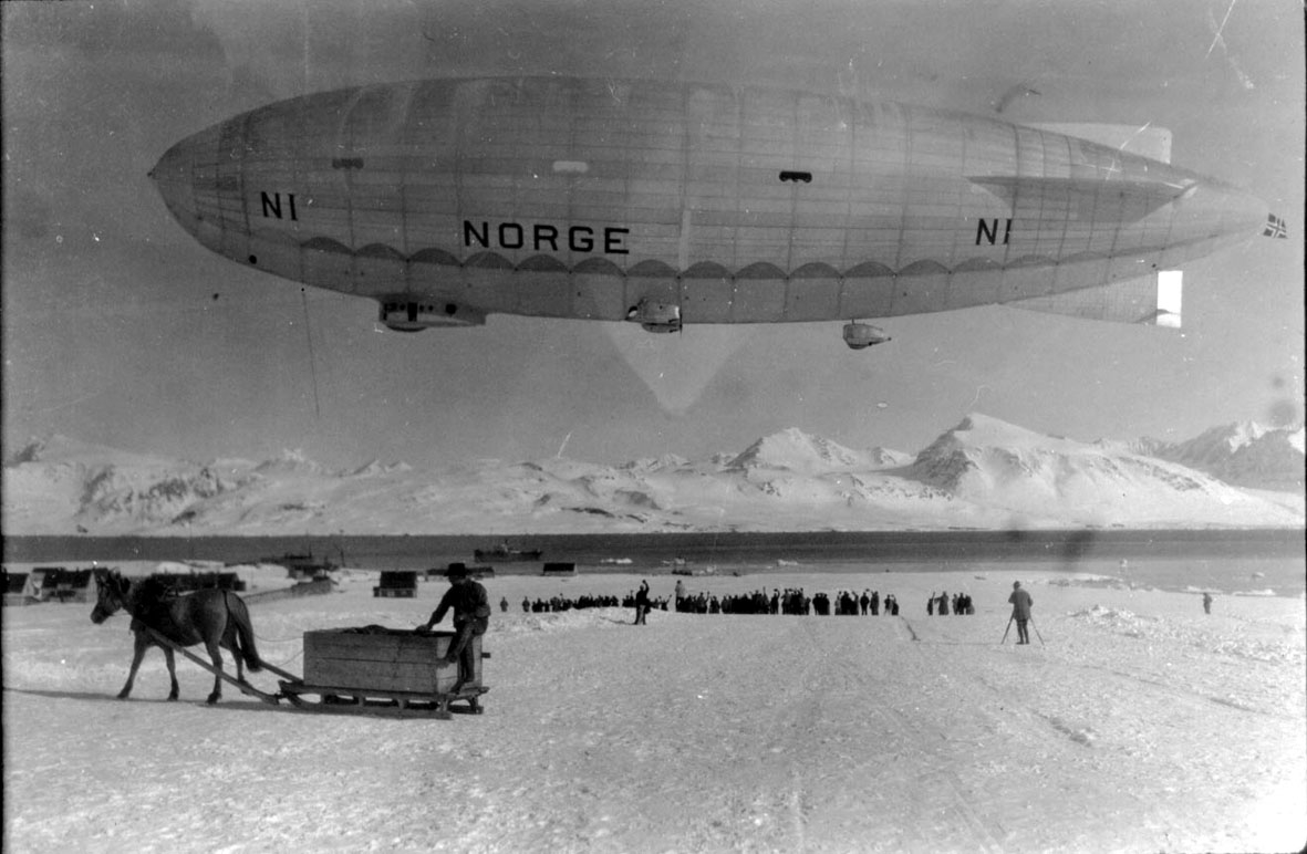 Luftskipet "Norge" like over bakken. Hest med slede i forgrunnen. Mange personer samlet nedenfor. Bak sees noen bygninger, havet med noen fartøy, fjell. Snø på bakken 