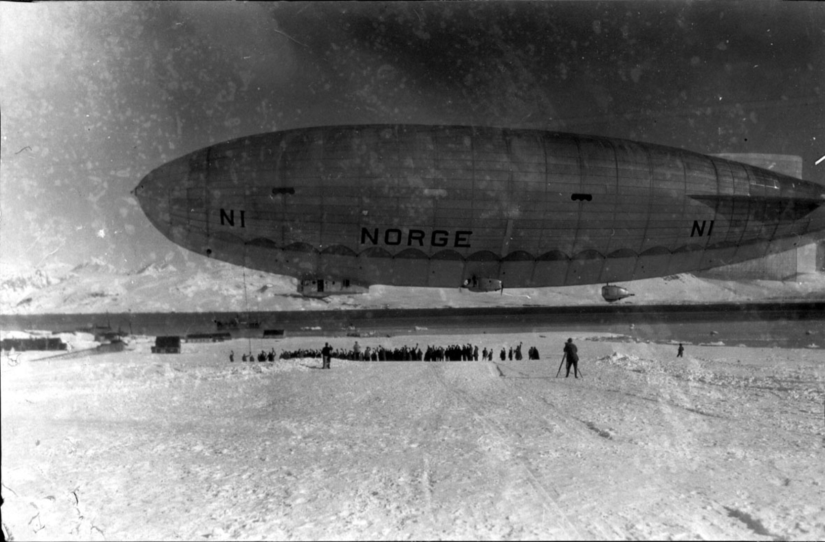 Luftskipet "Norge" like over bakken, letter med kurs for Nordpolen. Flere personer samlet under.
