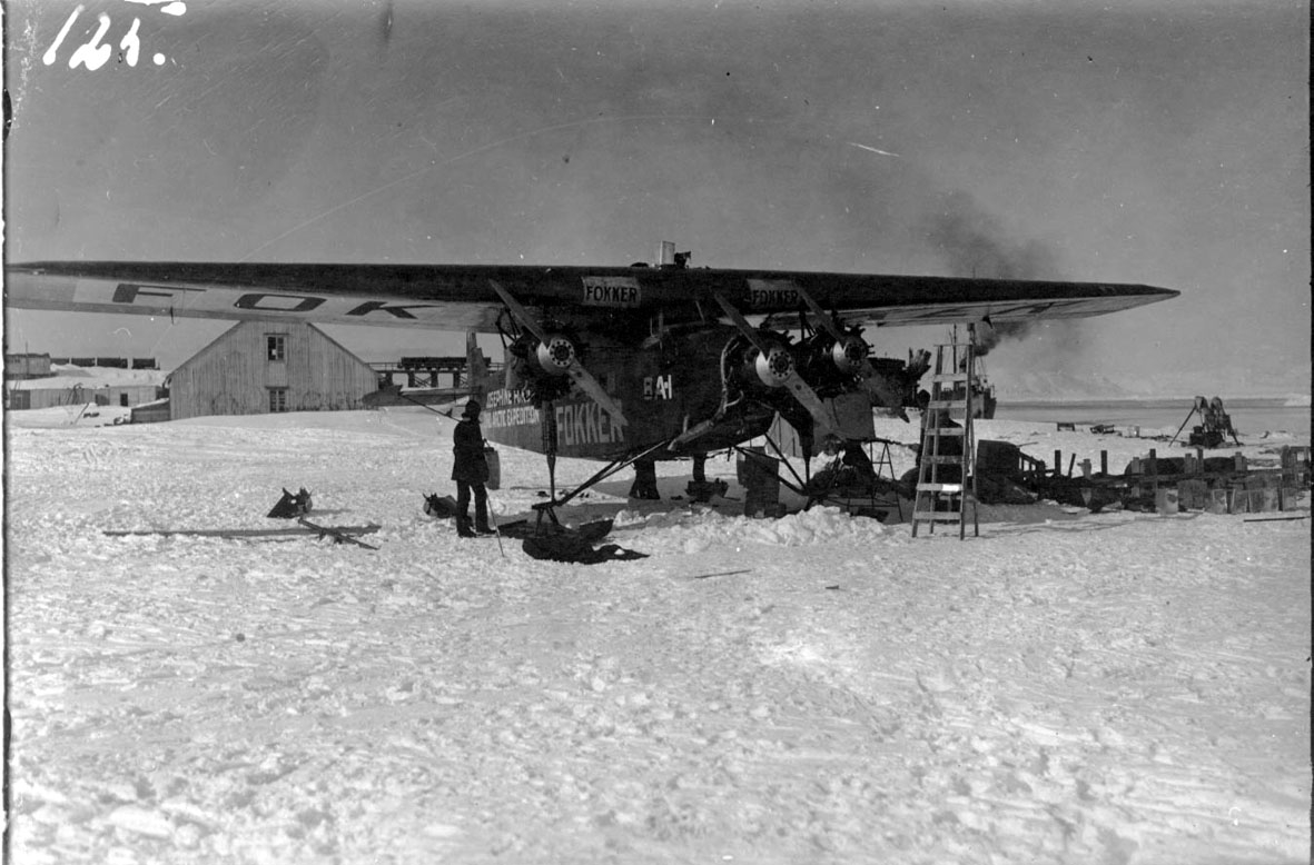 Fly, propellfly, på bakken, Fokker FVII "Josephine Ford". 1 person. Div. utstyr på bakken, stige. Bygning bak. Litt av havet t.h., snø på bakken.