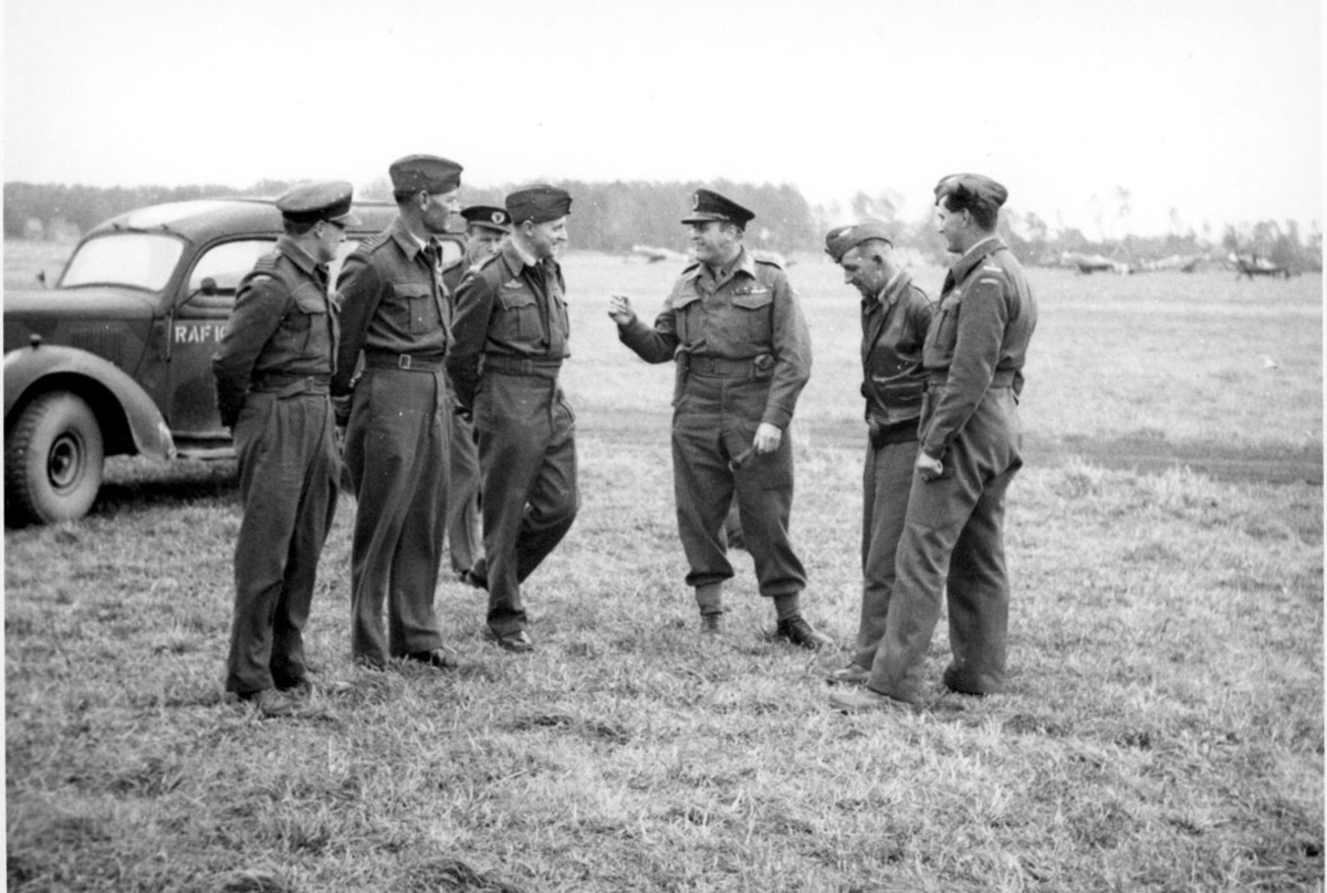 7 personer i militæruniform. Kronprins Olav og 6 andre,  på en åpen plass. Bil bak, med påskrift RAF, på siden. I noen fly i bakgrunnen.