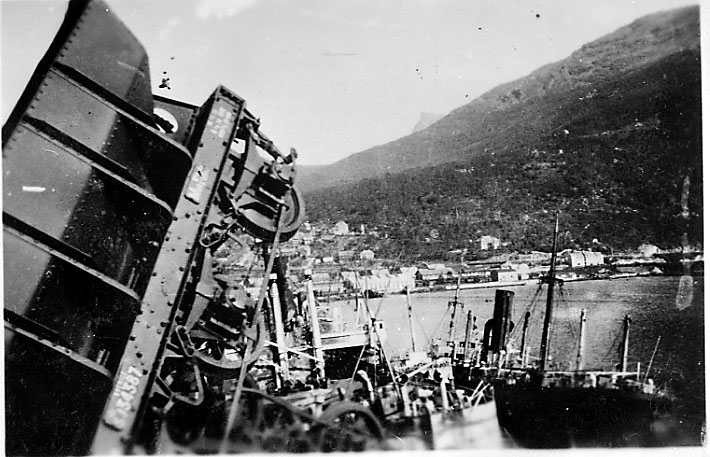 Nede ved havna, jernbanevogn ligger på siden, noen fartøy ved kai, bebyggelse. Narvik under 2. verdenskrig.
