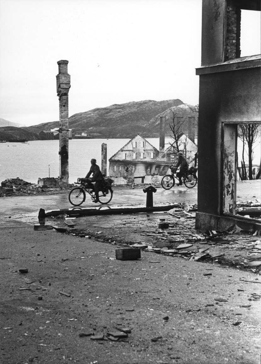 By - tettsted. Vei med to personer, menn i militæruniform på sykkel. Ruiner av flere bygninger. Hav - havneområde i bakgrunnen.
Tyske soldater i Sjøgata.