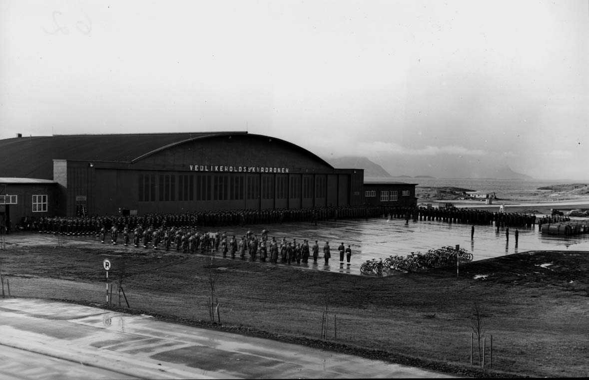 Lufthavn - flyplass. Militært personell. Oppstilling. Mange personer, menn, foran bygning - hangard merket vedlikeholdsskvadronen.