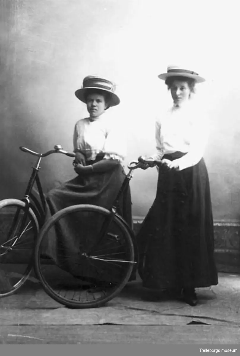 Två damer i vit blus, svart lång kjol, halmhatt och med cyklar. Negativ 70:3116.