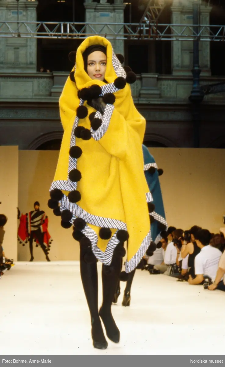 Modevisning. Modell i gul kappa med kapuschong och svarta bollar. Från Balmain.