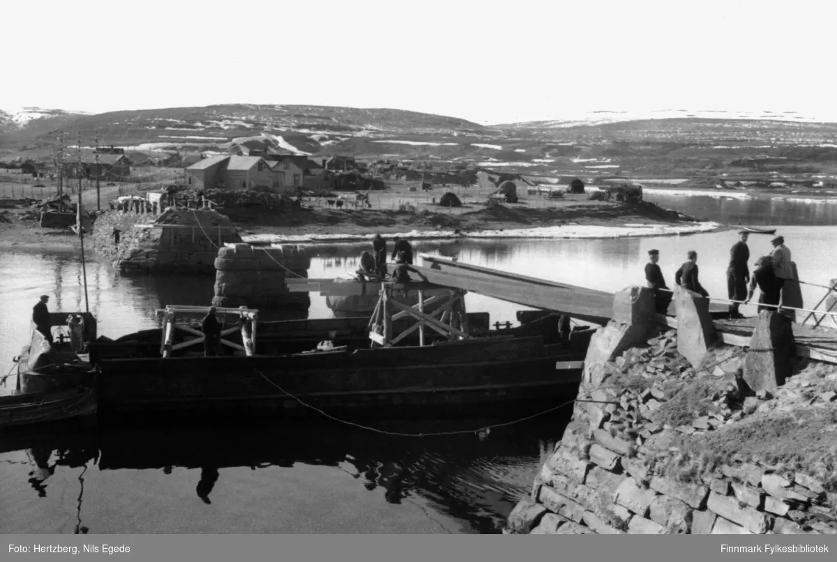 Bygging av Vestre Jakobselv bru, 1946. En båt ligger under den nylig påbegynte bruen. To menn er ombord i båten, fem menn jobber på bruen. Tre gutter står å ser på arbeidet. I bakgrunnen ser man noen hus.