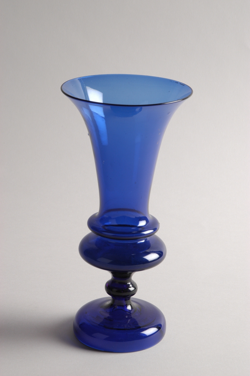 Vase (blomstervase) i mørkeblått, gjennomskinnelig glass. Blomsterkalkformet beger over fotstykke og profilert stett.