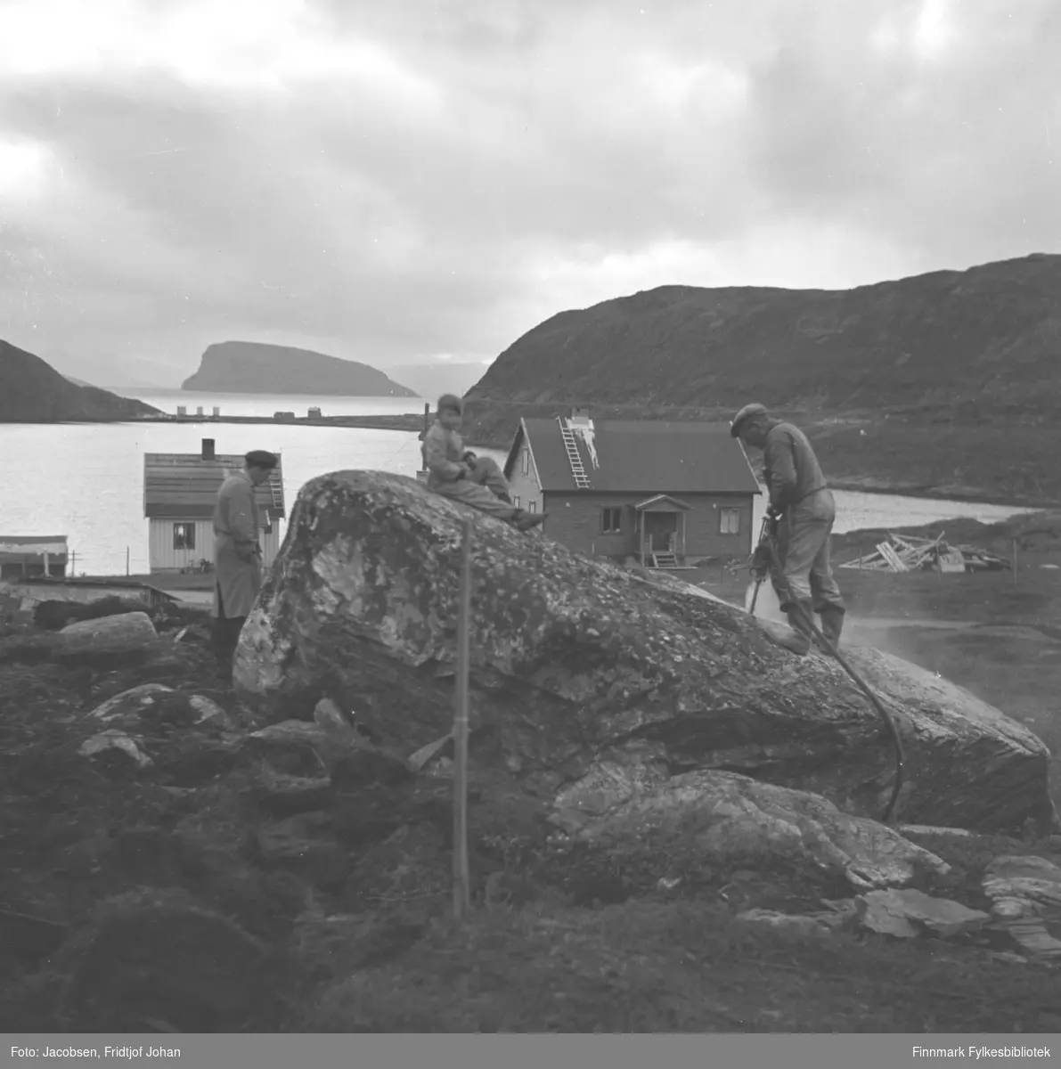 Arne Nakken står med trykkluftbor på en stor stein på eiendommen sin i Rypefjord. En gutt sitter oppå steinen og en mann står i lys frakk til venstre for steinen. To hus og taket på et tredje står litt lengre ned og en haug med materialer ligger i terrenget til høyre på bildet. Havna i Rypefjord  ses midt på bildet og fjellet til høyre er Rypefjell. Helt til høyre på bildet står Rypklubben med Rypklubbeidet mellom de to fjellene. Øya Håja ses i bakgrunnen, over Rypklubbeidet.