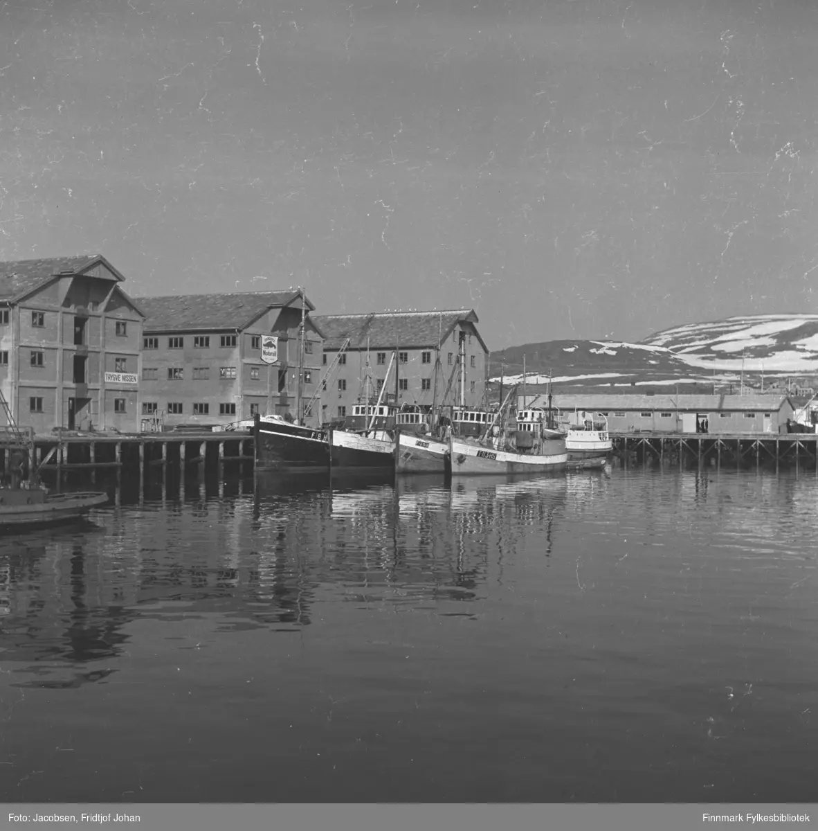 En vårdag i Hammerfest havn. De to byggene til venstre på bildet er pakkhusene til Trygve Nissen. Bygget ved siden av dem, midt på bildet, er Televerkets lager, Hammerfest folkebibliotek i dag. Den avlange brakka som står på kai til høyre på bildet er kailagret til FFR. Flere skøyter ligger fortøyd ved Nissen-kaia og hekken på en annen båt ses helt til venstre på bildet. Sykehuset kan ses rett over taket på FFR-brakka. Fjellet i bakgrunnen er Vardfjell med Storfjellet til høyre. En del snø ligger i fjellet enda som som vinter om en fin vårdag.