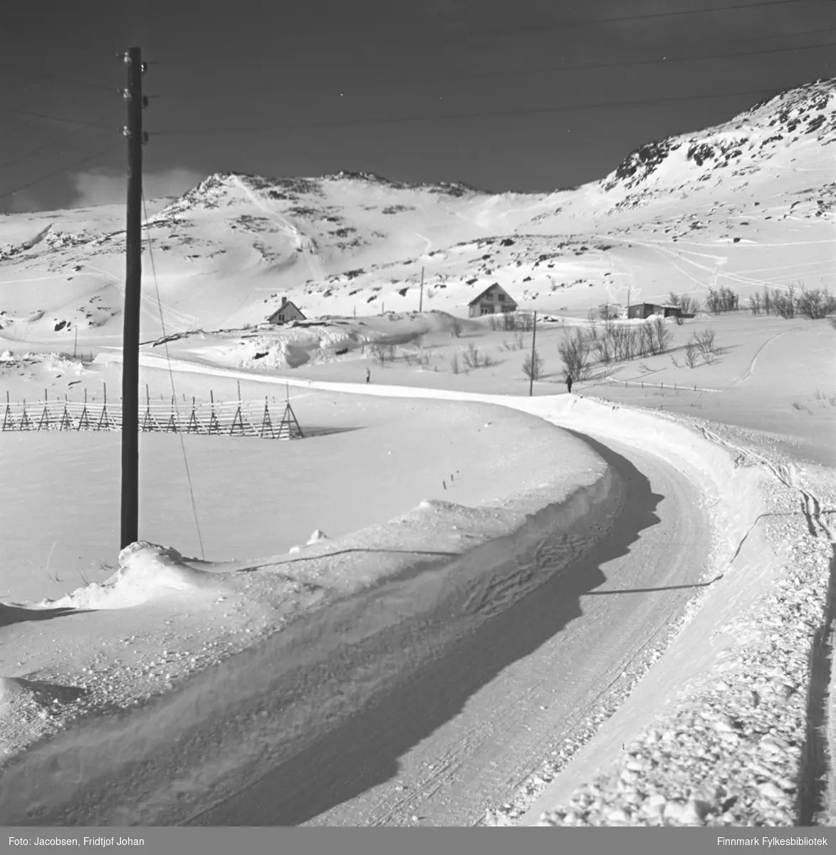 Motiv fra Storneset i Rypefjord langs en brøytet vei i retning Kubakken og Hammerfest. Noen bygninger står litt bak i terrenget. Til venstre står en lang snøskjerm og en el-stolpe. To personer går på ski langs veikanten og skispor ses over deler av terrenget.