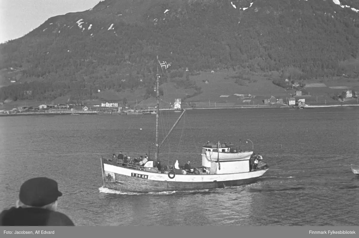 Bilde av en motorskøyta T 34 KN "Segelvik" , antagelig avbildet i  Tromsøysundet. Eierne av båten var Olaf Johnsen, Emil Johnsen og Markus Berg. Segelvik var en av Nygaardsvoldbåtene. Båten er bygget av tre. Rorhuset står akterut, er hvitmalt og har en livbåt hengende på siden. En person står på hekken. På baugen står flere personer. Rett ved siden av dem står masta. Den har det norske flagg i toppen og bommen stikker ut ganske langt nede på masta. Et ganske høyt fjell står i bakgrunnen, delvis skogkledt. Noen bygninger står i strandkanten og en kai ses til til venstre på bildet. Helt nederst er en mann med mørk overdel og mørk skyggelue som står og ser på båten. Sjøen ligger svært rolig og det ser ut til å være flott vær en sen vårdag eller tidlig sommerdag.