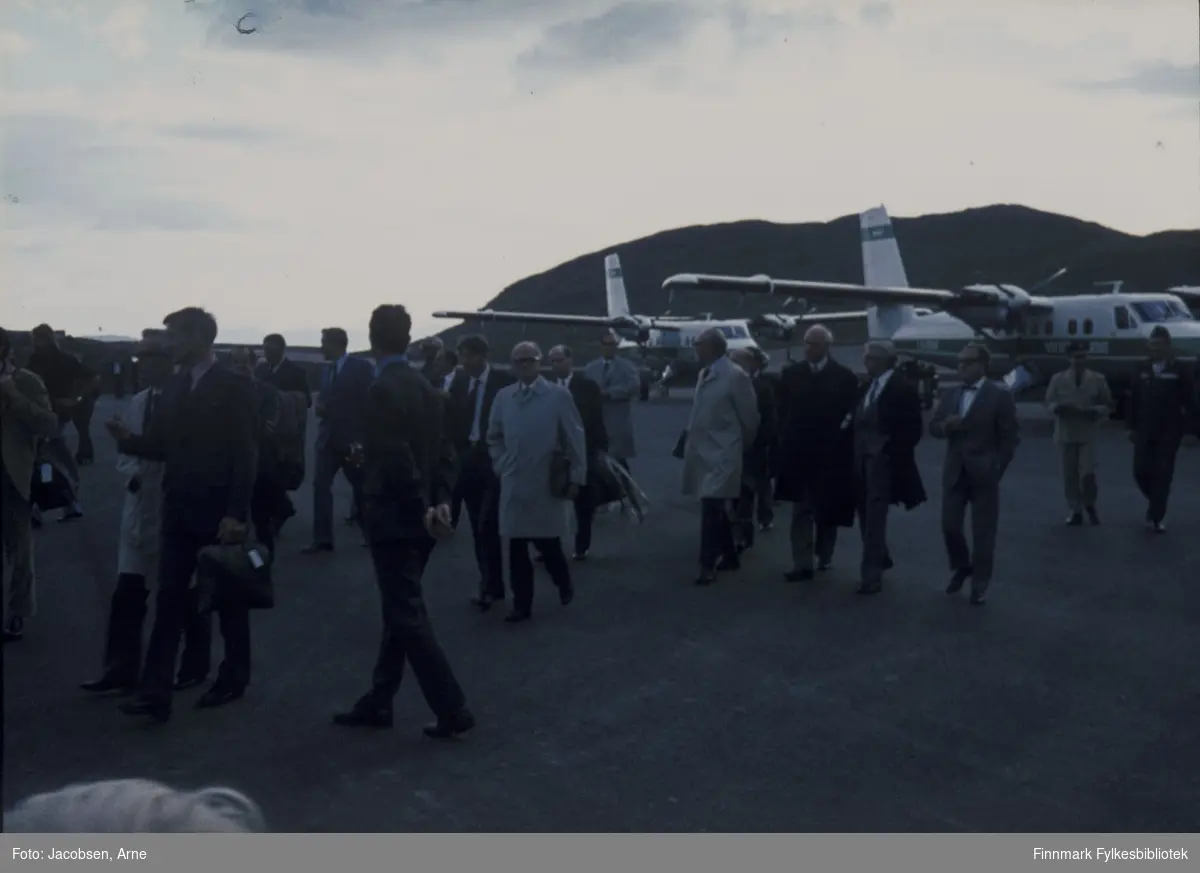 To Twin Otter-fly fra Widerøes flyveselskap parkert på Hammerfest lufthavn, med snuten mot Reindalen, under åpningen 30. juli 1974. Mange, pent kledde mennesker går rundt på flyplassen for å se og delta på åpningen. De går mot terminalbygningen og tårnet så visningen av flyene er nok over. Bak flyene ses fjellet Vardfjell.