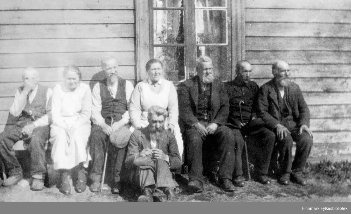Fotografi av ukjente personer. To kvinner og seks eldre menn. De sitter på en benk med ryggen til et trehus. Det er et vindu med hvite gardiner og blomster, på veggen. En mann sitter foran på bakken. De fleste av mennene har skjegg, og er mørkkledte. To av dem har stokk. Kvinnene har hvite forklekjoler på seg