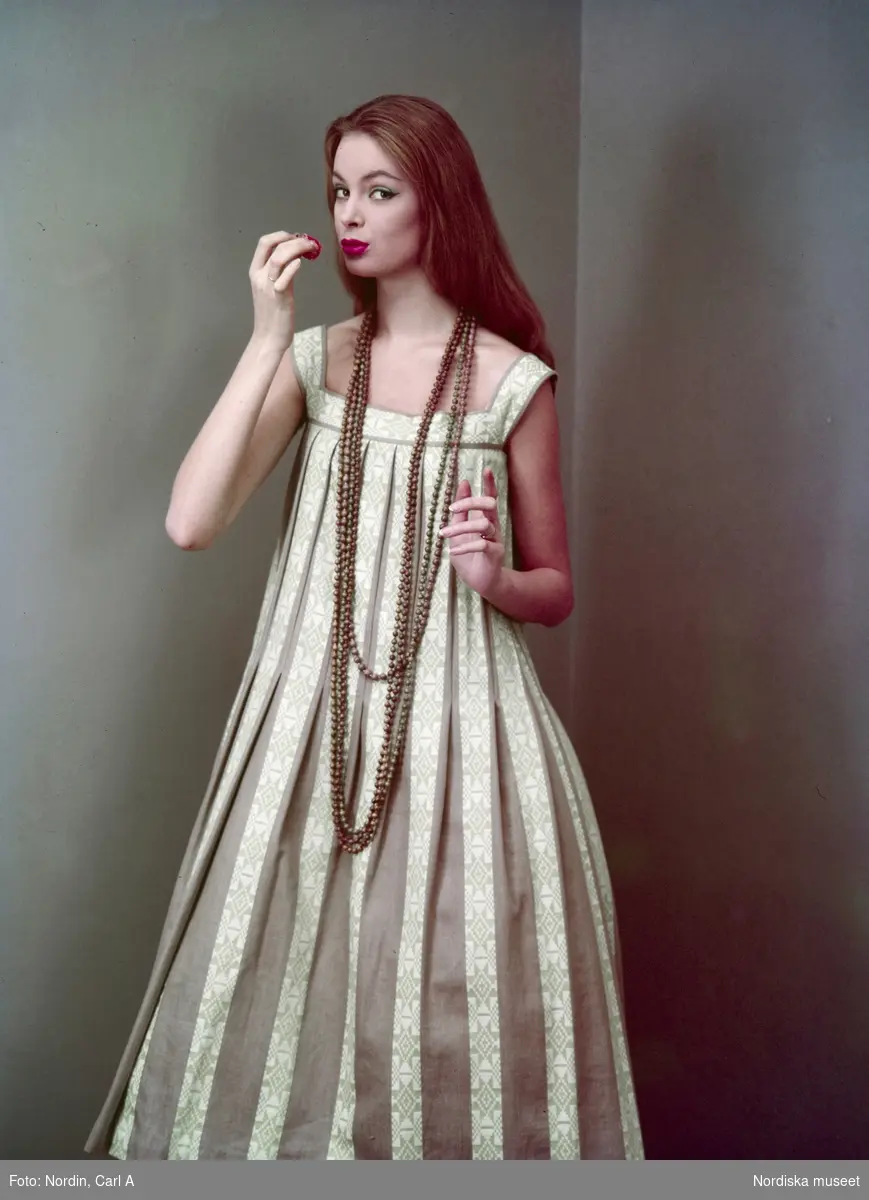 Modell i randig klänning med mönster, halsband och långt, rött hår med jordgubbe i handen.