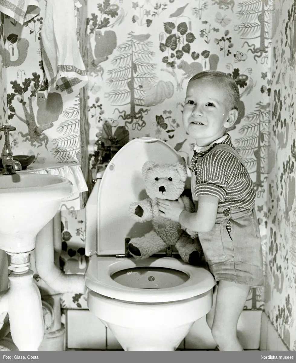 Ett barn i randig tröja och hängslebyxor med nalle i handen, vid en toalett.