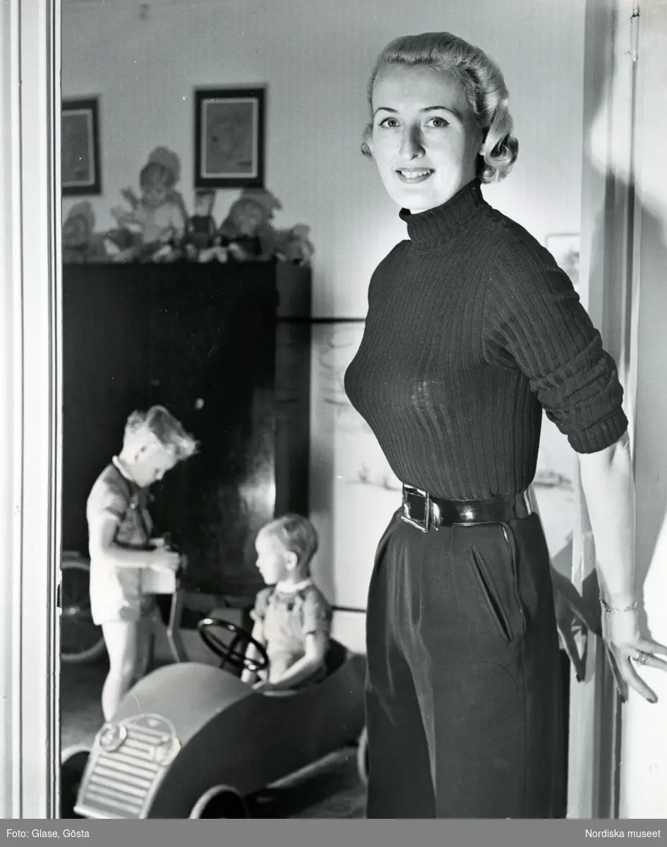 Kvinna, mannekängen Kim Söderlund klädd i polotröja, byxor och lackskärp. Två barn leker med leksaksbil i bakgrunden.