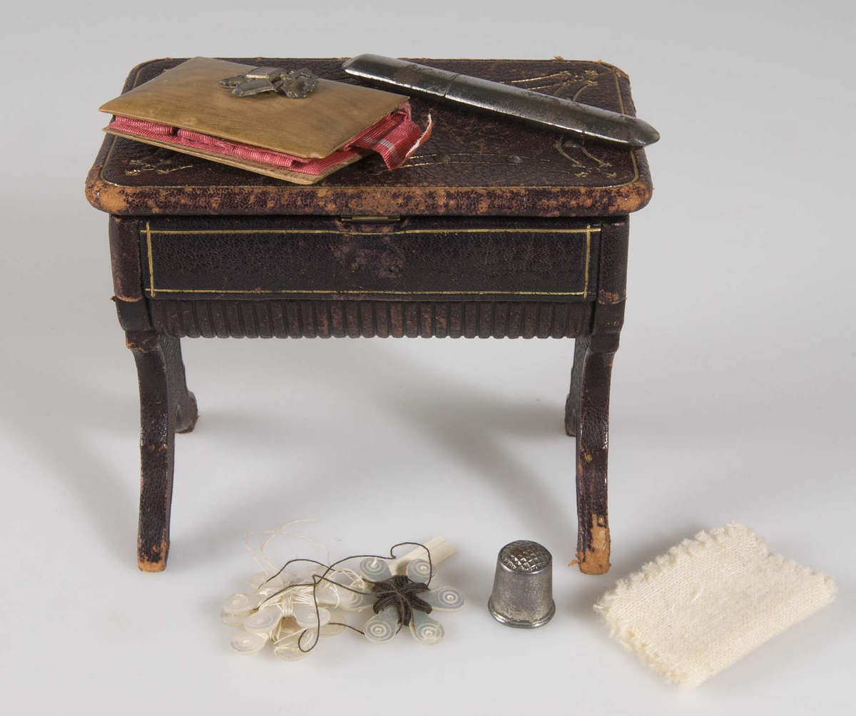 Litet sybord för dockskåp i brunt läder med innehåll: tre trådnystan, en fingerborg i metall, en bok med träpärmar, ett fodral i metall samt två vita tygbitar.