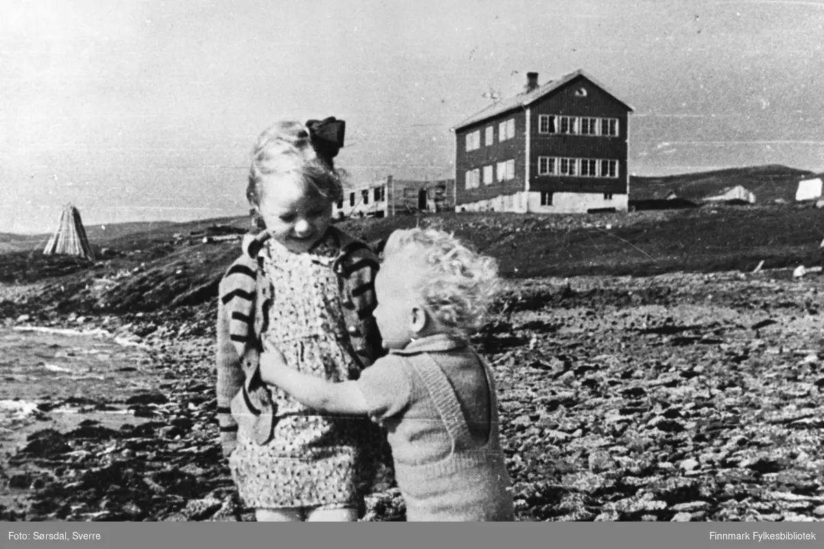 Bildet viser to barn som leker i fjæra i Hamna i Syltefjord. Barna som leker er Randi og Eirik Sørsdal. I bakgrunnen ser man skoleinternatet. Vardø sykehus ble flyttet hit etter bombingen i Vardø under 2. verdenskrig. Ved siden av skoleinternatet bygges det bolig for de ansatte, med materialer fra bombede hus i Vardø. Bildet er tatt høsten 1942.