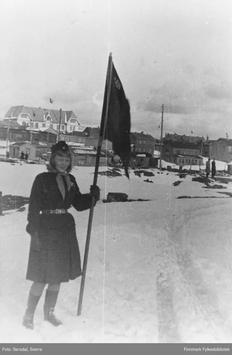 Bilde av Randi Sørsdal i speideruniform som holder speiderflagget. Det er 17. mai 1952. I bakgrunnen ses skolen i Vardø.