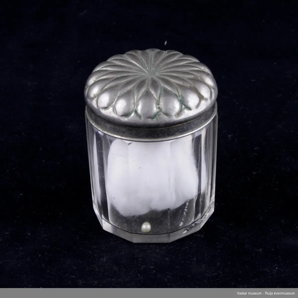 Glassbeholder med lokk av sølvplett. Inni finnes en bomullsdott og en liten perle.