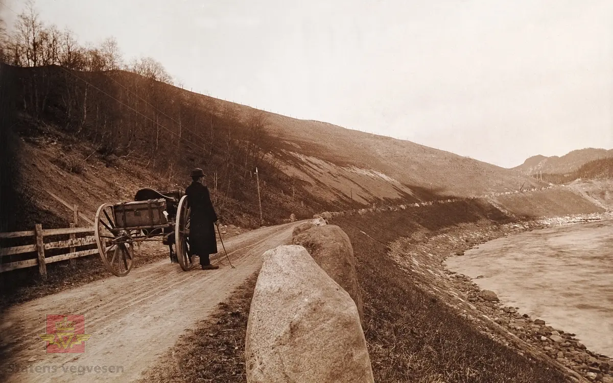"Veianlæg". "Langs Orkla". Mann ikledd frakk og hatt står ved hest  med reisekoffert- amerikakoffert på karjolen. Bildet kan være tatt før år 1900.