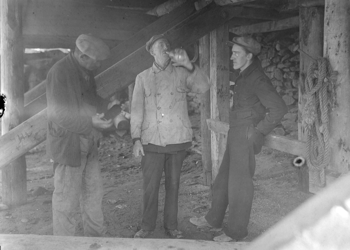 Tre menn drikker brennevin under ei brygge