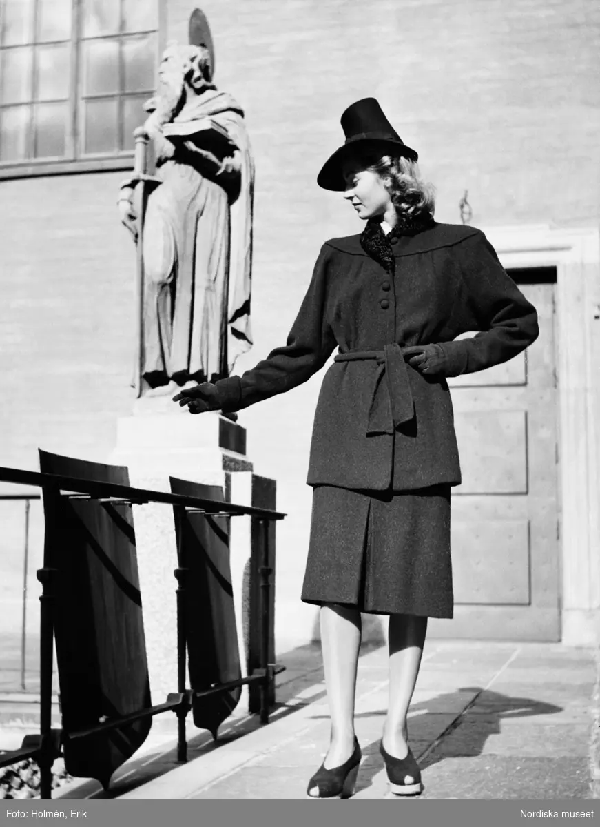 Nordiska Kompaniet visar dammode. Modell klädd i mörk dräkt med skärp och klädda knappar, samt hatt och handskar. Skor med öppen tå och kilklack. I bakgrunden syns en staty.