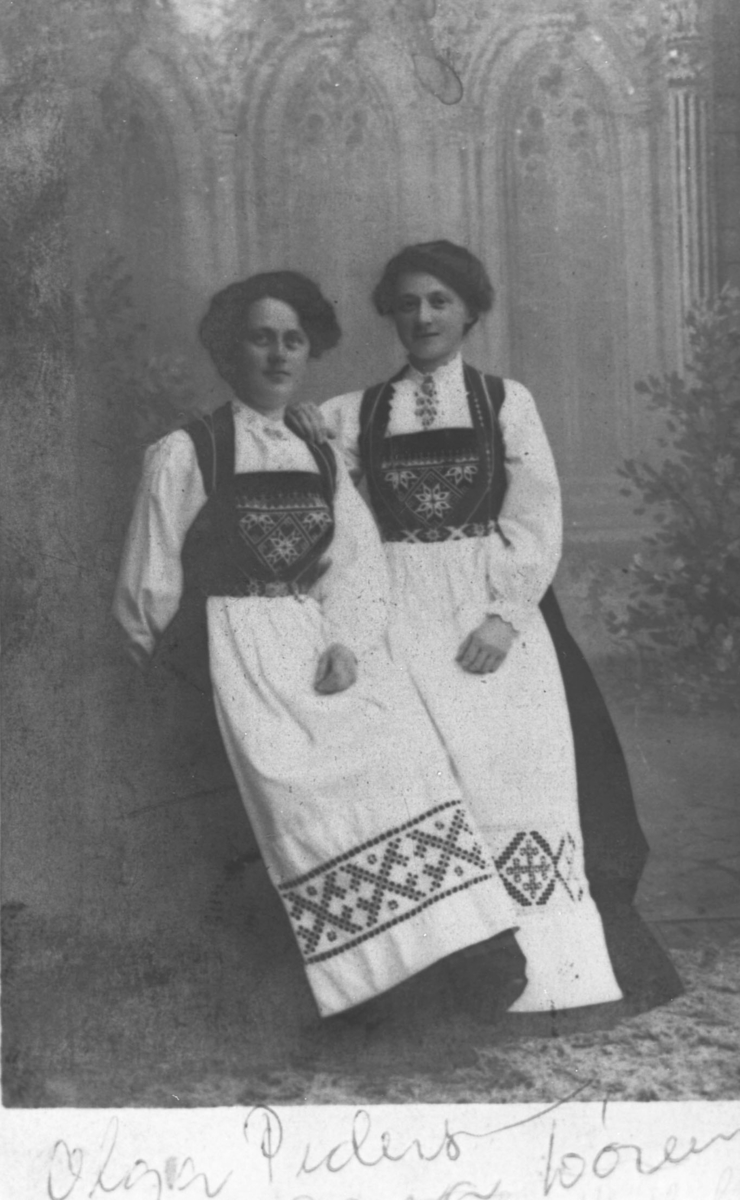 Kabinettportrett av to unge kvinner i bunader. Fra venstre: Olga Marie Pedersen og Lise Hendriksen. Disse unge damene er ca 16-20 år gamle. Bildet er opprinnelig kopiert til postkortform (postkortmarkering på baksiden) men senere klep til og passe in i visitkortalbumet.