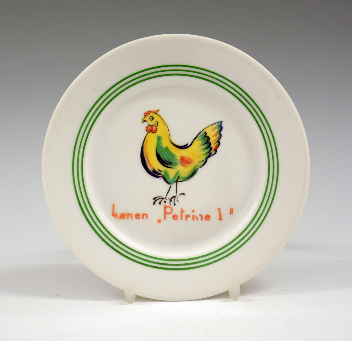 Asjett i porselen til dukkeservise. Hvit glasur. Dekorert med høne og tekst hønen Petrine I. Trykkdekor, grønn strek.
Modell: 1460.
Dekor: "Hønen Petrine 1".