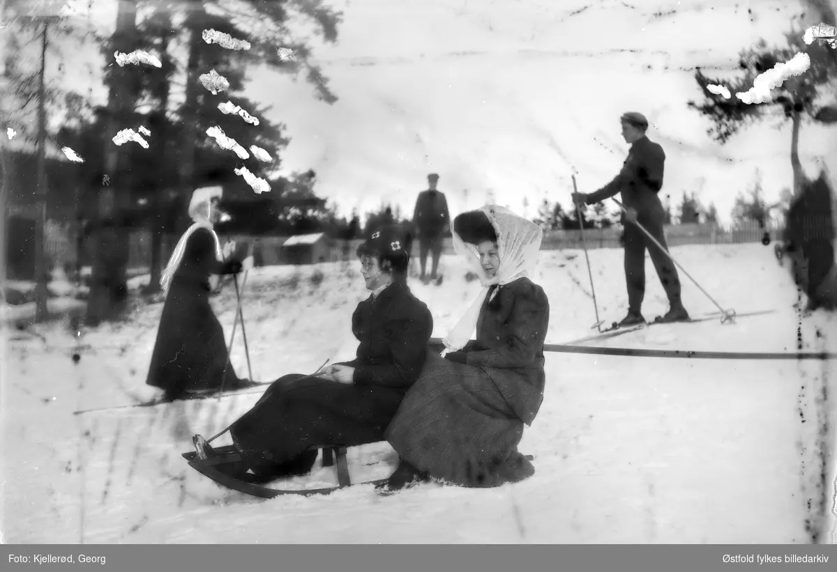 To kvinner på kjelke, skiløpere i bakgrunnen. Antakelig Nordmarka i Oslo.
