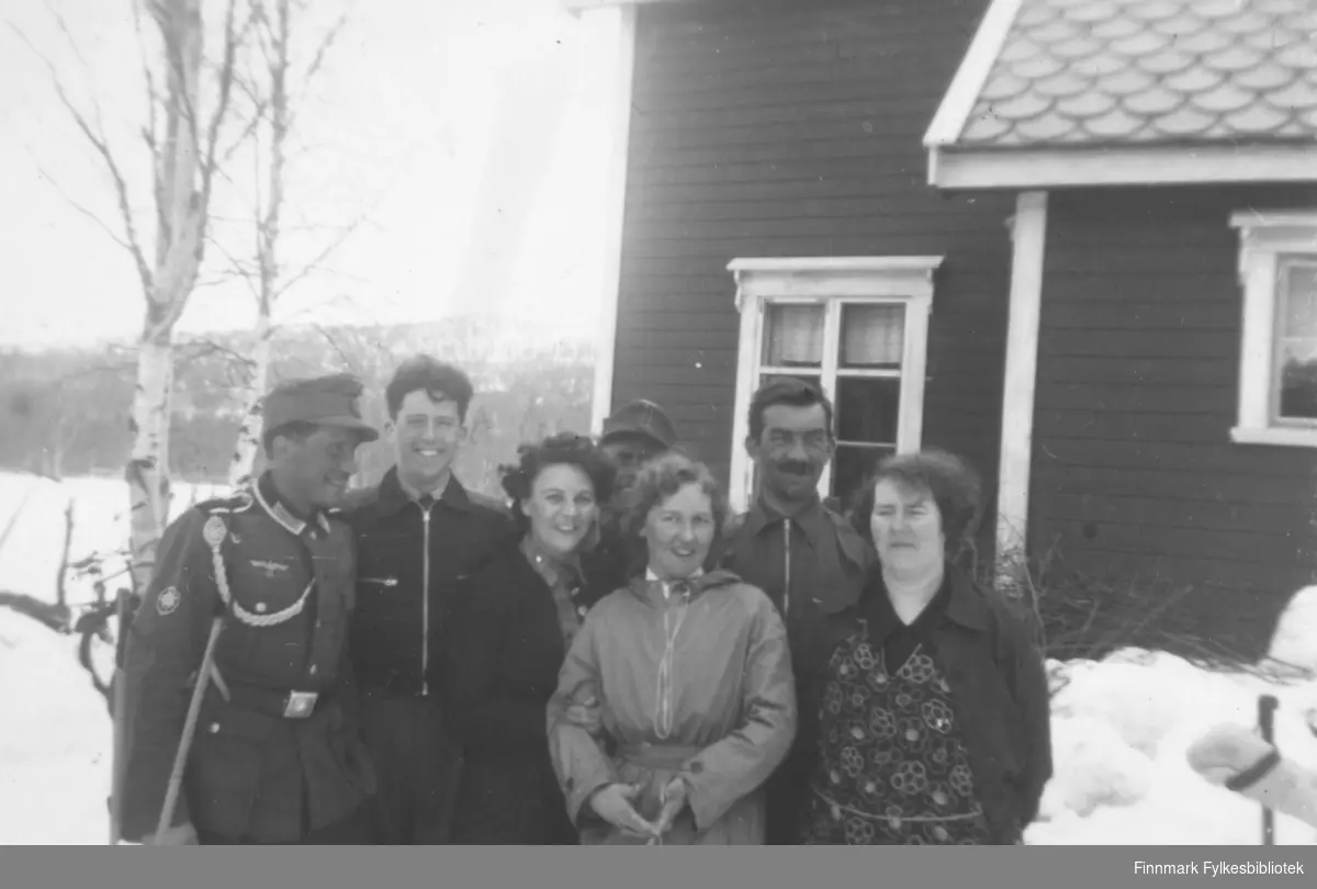 Altafolk i Gargiadalen under 2. verdenskrig. Fire menn og tre kvinner. To av mennene er uniformskledd. På sidene ser vi noen skistaver stukket ned i snøen og i bakgrunnen trær og et bolighus. Ca.1935-1945.