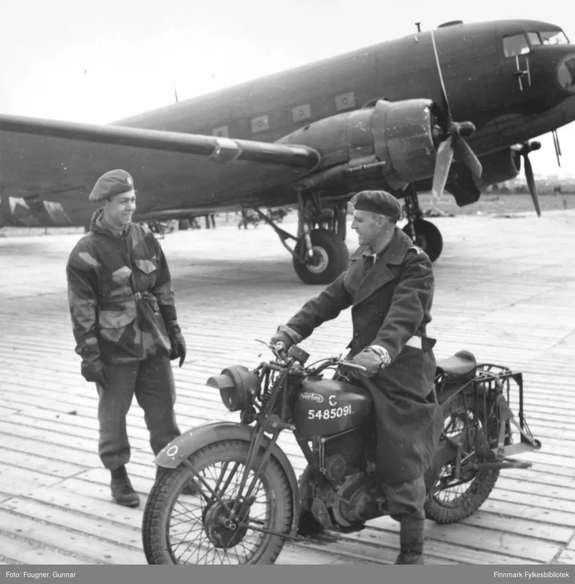 På Banak flyplass. En soldat og en offiser i uniformer. Offiseren sitter på en 'Norton' motorsykkel. I bakgrunnen står et militært fly av typen C-47, Dakota.