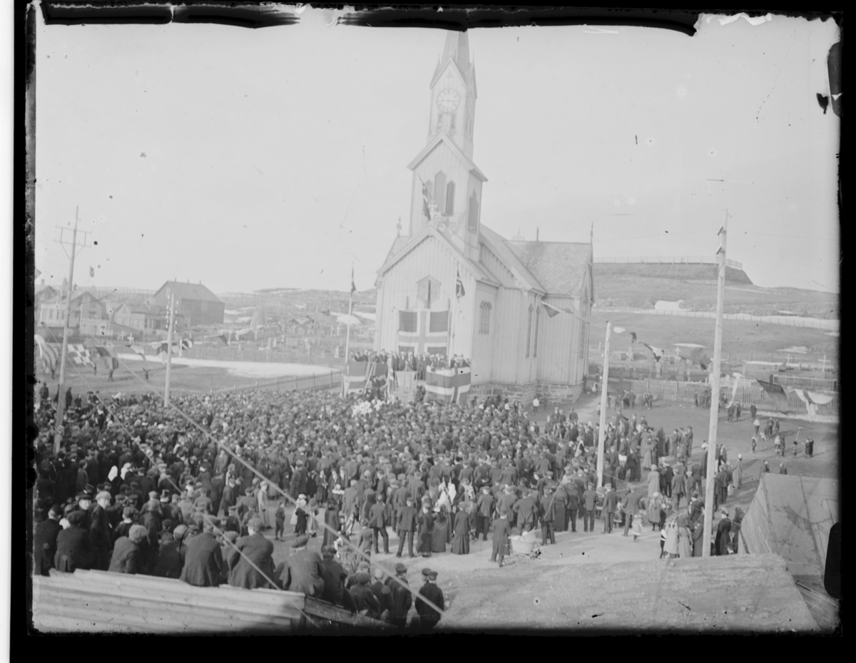 Dette kan være en 17.mai-feiring i Vardø en stor folkemengde samlet utenfor kirka. På kirketrappa er det laget talerstol og pyntet med flagg. Bak kirka ser vi deler av kirkegården og Klondyke. I forgrunnen ser vi remser med signalflagg hengt opp som utsmykning